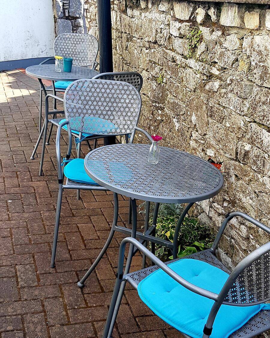 Café tables and chairs with turquoise cushions on a paved courtyard beside a stone wall